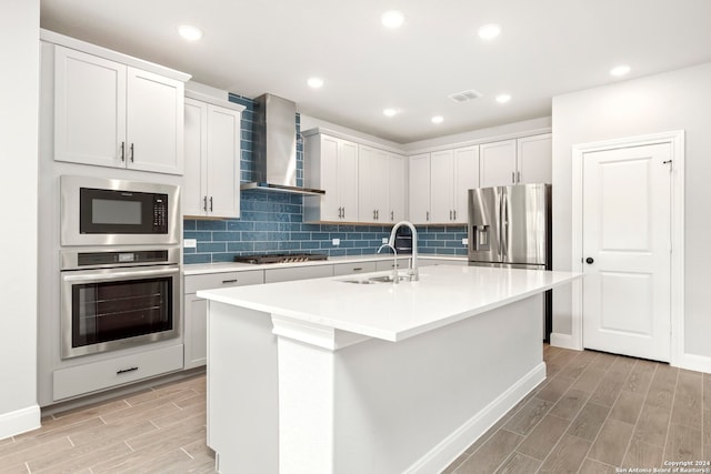 kitchen featuring stainless steel appliances, sink, wall chimney range hood, a center island with sink, and white cabinets