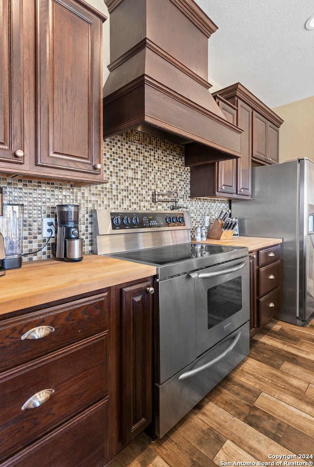 kitchen with decorative backsplash, dark hardwood / wood-style flooring, premium range hood, butcher block counters, and stainless steel appliances