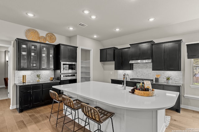 kitchen featuring light wood-type flooring, a kitchen bar, an island with sink, backsplash, and stainless steel appliances