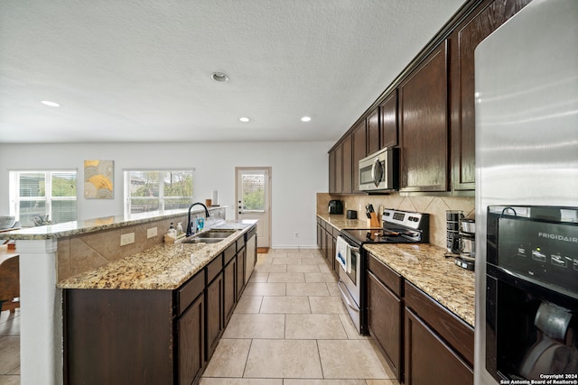 kitchen featuring a kitchen island with sink, sink, dark brown cabinetry, appliances with stainless steel finishes, and light stone counters