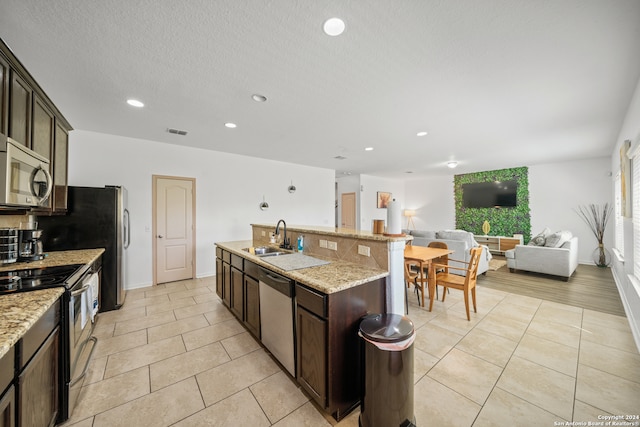 kitchen with backsplash, appliances with stainless steel finishes, a kitchen island with sink, light stone countertops, and dark brown cabinetry