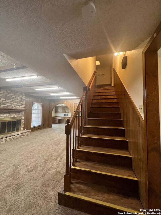 staircase featuring wood walls, a stone fireplace, a textured ceiling, and carpet flooring