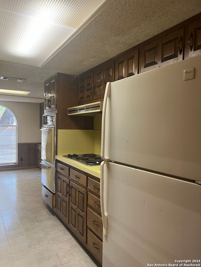 kitchen with exhaust hood, a textured ceiling, wood walls, dark brown cabinetry, and white appliances