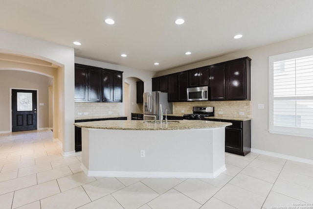 kitchen with tasteful backsplash, light stone counters, appliances with stainless steel finishes, light tile patterned floors, and an island with sink