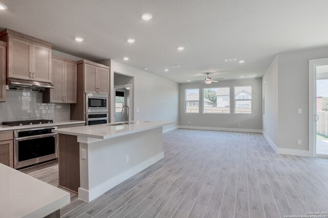 kitchen with appliances with stainless steel finishes, decorative backsplash, an island with sink, light hardwood / wood-style floors, and ceiling fan