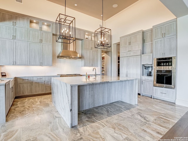 kitchen featuring wall chimney range hood, pendant lighting, light stone countertops, an island with sink, and stainless steel double oven