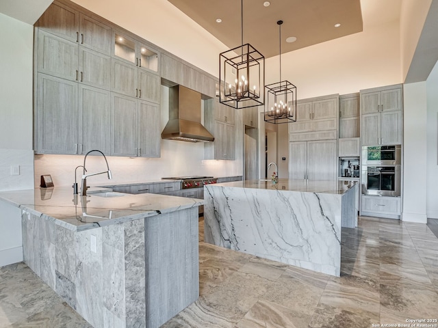 kitchen featuring wall chimney range hood, pendant lighting, sink, a kitchen island with sink, and light stone countertops