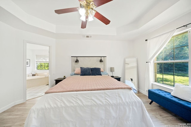 bedroom with a raised ceiling, connected bathroom, ceiling fan, and light wood-type flooring