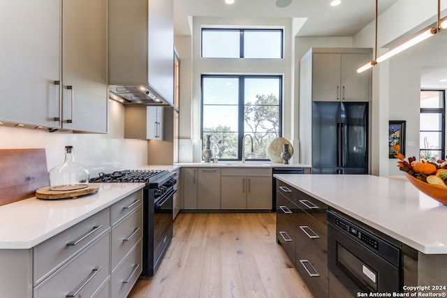 kitchen featuring wall chimney exhaust hood, hanging light fixtures, sink, black appliances, and light hardwood / wood-style floors