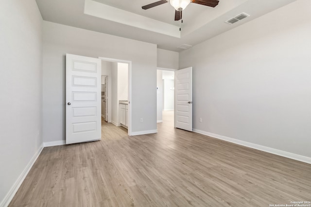 unfurnished bedroom featuring ceiling fan, ensuite bath, light wood-type flooring, and a tray ceiling