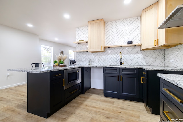 kitchen with wall chimney range hood, kitchen peninsula, stainless steel microwave, light hardwood / wood-style flooring, and sink