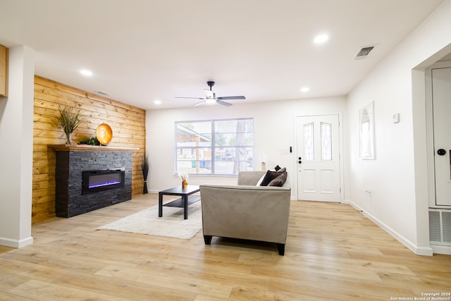 living room featuring ceiling fan, wood walls, light wood-type flooring, and a stone fireplace