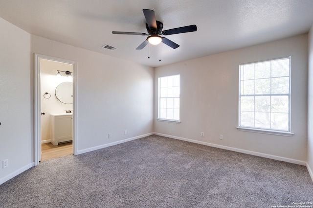 unfurnished bedroom featuring multiple windows, ensuite bath, light colored carpet, and ceiling fan