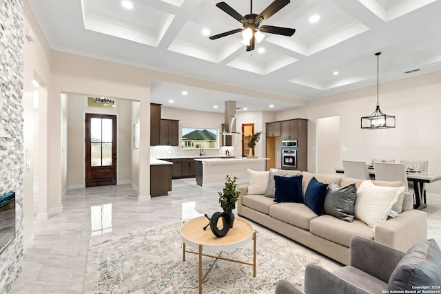 living room with a fireplace, ceiling fan with notable chandelier, coffered ceiling, beamed ceiling, and crown molding