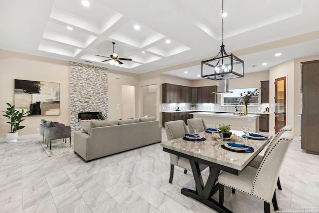 dining area with beam ceiling, coffered ceiling, a stone fireplace, and ceiling fan with notable chandelier