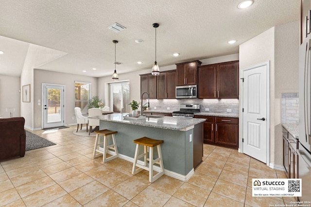 kitchen featuring a kitchen island with sink, hanging light fixtures, stainless steel appliances, backsplash, and light stone counters