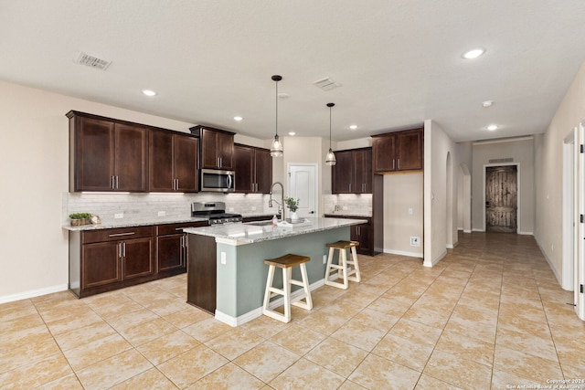 kitchen with light stone countertops, appliances with stainless steel finishes, backsplash, decorative light fixtures, and a kitchen island with sink