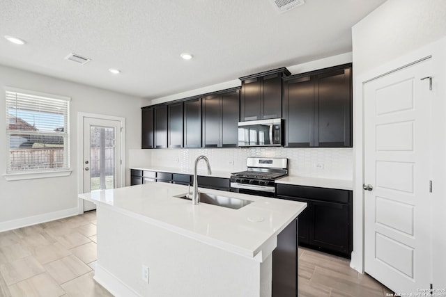 kitchen with sink, a textured ceiling, stainless steel appliances, a kitchen island with sink, and decorative backsplash