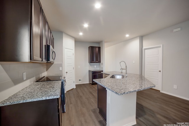 kitchen featuring a center island with sink, dark brown cabinets, a sink, stainless steel microwave, and dark wood-style floors