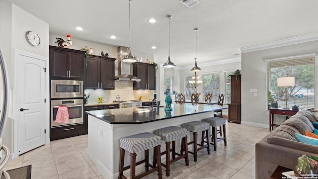 kitchen with wall chimney range hood, hanging light fixtures, appliances with stainless steel finishes, a kitchen island with sink, and ornamental molding