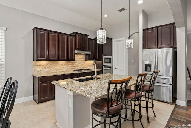 kitchen with hanging light fixtures, stainless steel appliances, a center island with sink, sink, and light stone counters