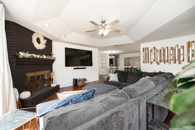 living room featuring hardwood / wood-style flooring, ceiling fan with notable chandelier, a raised ceiling, and a brick fireplace