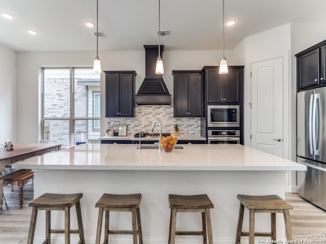 kitchen with appliances with stainless steel finishes, custom range hood, decorative light fixtures, and an island with sink