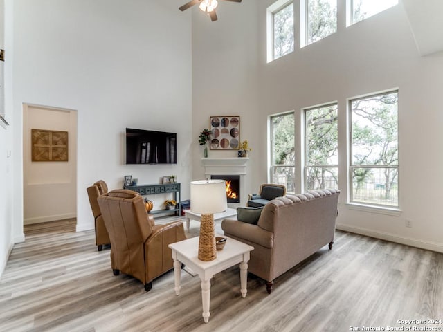 living room featuring a towering ceiling, a lit fireplace, light wood finished floors, ceiling fan, and baseboards