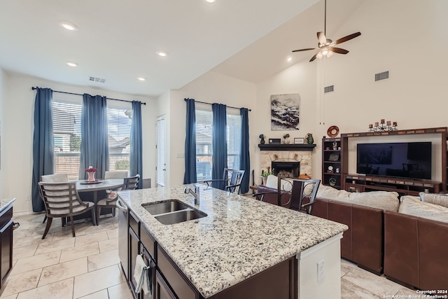 kitchen featuring ceiling fan, a kitchen island with sink, sink, a fireplace, and light stone counters