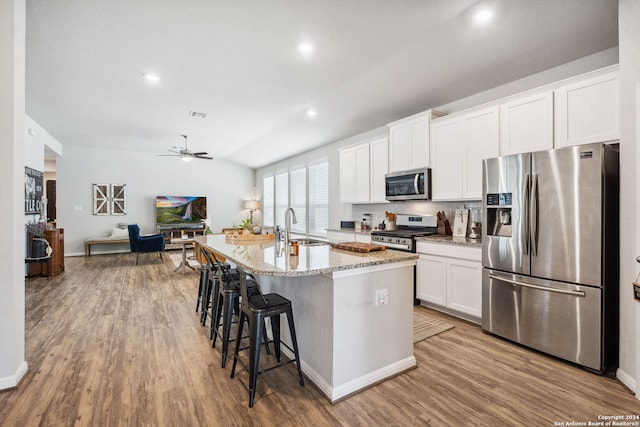 kitchen featuring appliances with stainless steel finishes, light hardwood / wood-style floors, white cabinets, light stone counters, and a kitchen island with sink