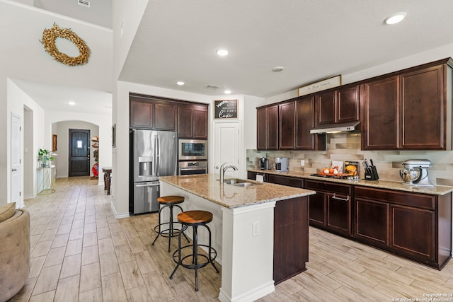 kitchen with a kitchen island with sink, stainless steel appliances, a breakfast bar, light wood-type flooring, and light stone counters