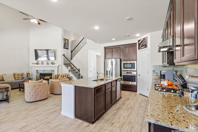 kitchen with dark brown cabinets, a center island with sink, extractor fan, light stone countertops, and stainless steel appliances