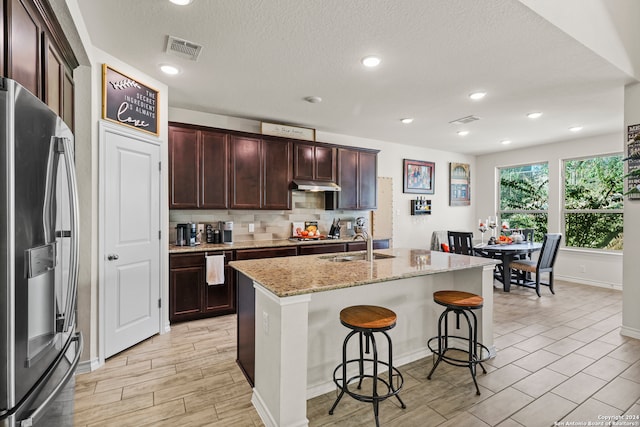 kitchen featuring stainless steel fridge, an island with sink, a breakfast bar, light stone countertops, and sink