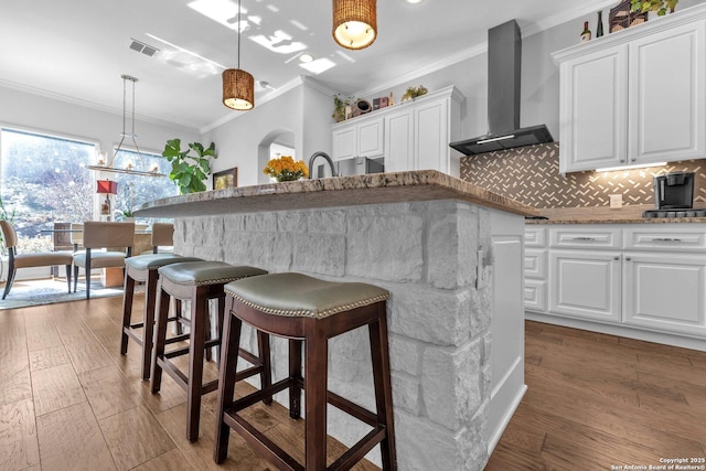 kitchen featuring pendant lighting, white cabinets, wall chimney range hood, ornamental molding, and a center island with sink