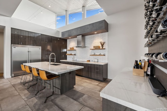 kitchen with sink, a kitchen bar, built in fridge, wall chimney exhaust hood, and a center island with sink