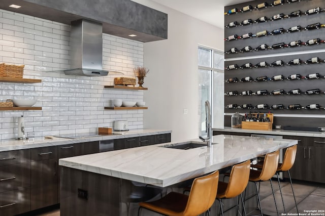 interior space with a kitchen breakfast bar, wall chimney exhaust hood, light stone counters, decorative backsplash, and a center island with sink