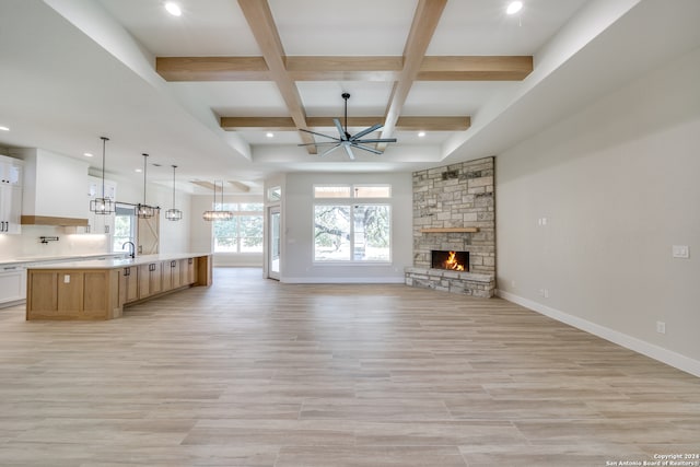 unfurnished living room with beam ceiling, a stone fireplace, ceiling fan with notable chandelier, and light hardwood / wood-style floors