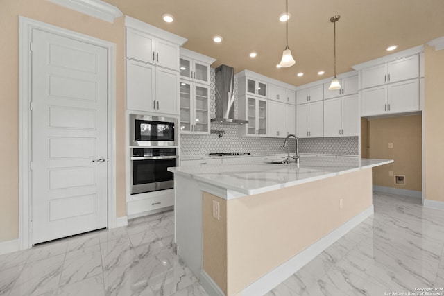 kitchen with wall chimney range hood, white cabinets, backsplash, an island with sink, and pendant lighting