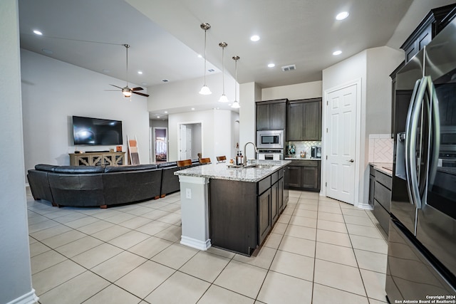 kitchen featuring an island with sink, stainless steel appliances, backsplash, dark brown cabinetry, and decorative light fixtures