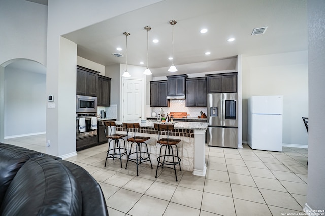 kitchen with a kitchen island with sink, hanging light fixtures, stainless steel appliances, backsplash, and dark brown cabinetry