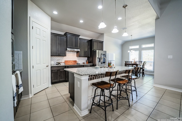 kitchen featuring light tile patterned floors, stainless steel fridge, decorative light fixtures, a breakfast bar, and a kitchen island with sink