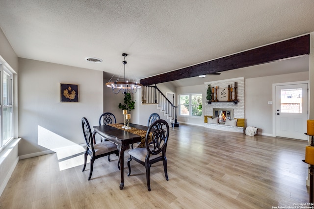 dining room featuring a chandelier, a textured ceiling, a brick fireplace, light hardwood / wood-style flooring, and beamed ceiling