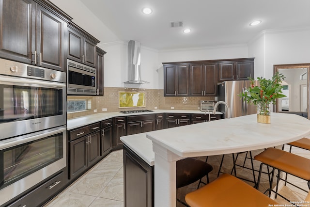 kitchen featuring a center island with sink, appliances with stainless steel finishes, wall chimney exhaust hood, a breakfast bar area, and crown molding