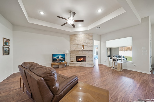 living room with ceiling fan, a stone fireplace, hardwood / wood-style flooring, and a raised ceiling