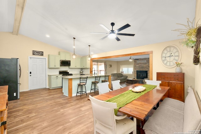 dining space with sink, vaulted ceiling with beams, light wood-type flooring, a stone fireplace, and ceiling fan