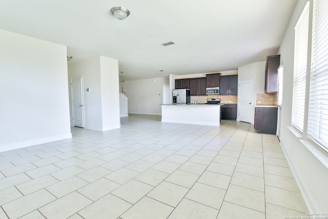 kitchen with dark brown cabinets, stainless steel appliances, a center island with sink, light tile patterned floors, and tasteful backsplash