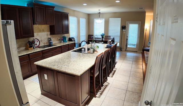 kitchen featuring hanging light fixtures, a center island with sink, sink, dark brown cabinetry, and light stone counters