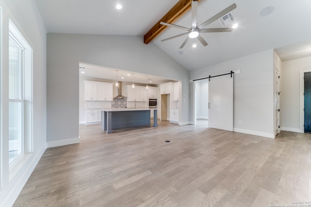 unfurnished living room featuring ceiling fan, beam ceiling, a barn door, and light wood-type flooring