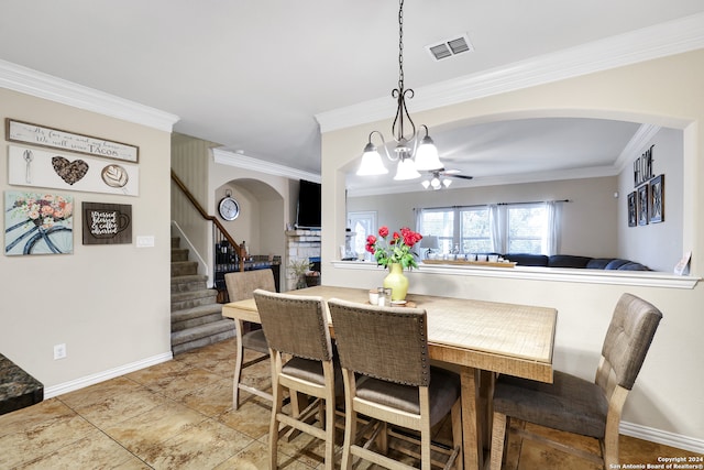 dining area featuring a stone fireplace, tile patterned flooring, ornamental molding, and ceiling fan with notable chandelier