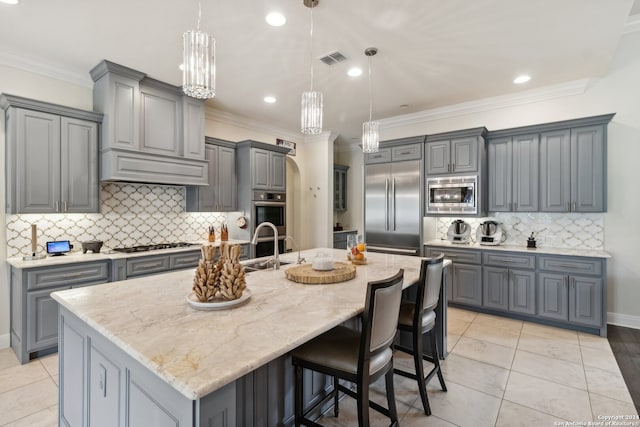 kitchen featuring a large island with sink, built in appliances, gray cabinets, pendant lighting, and light stone countertops